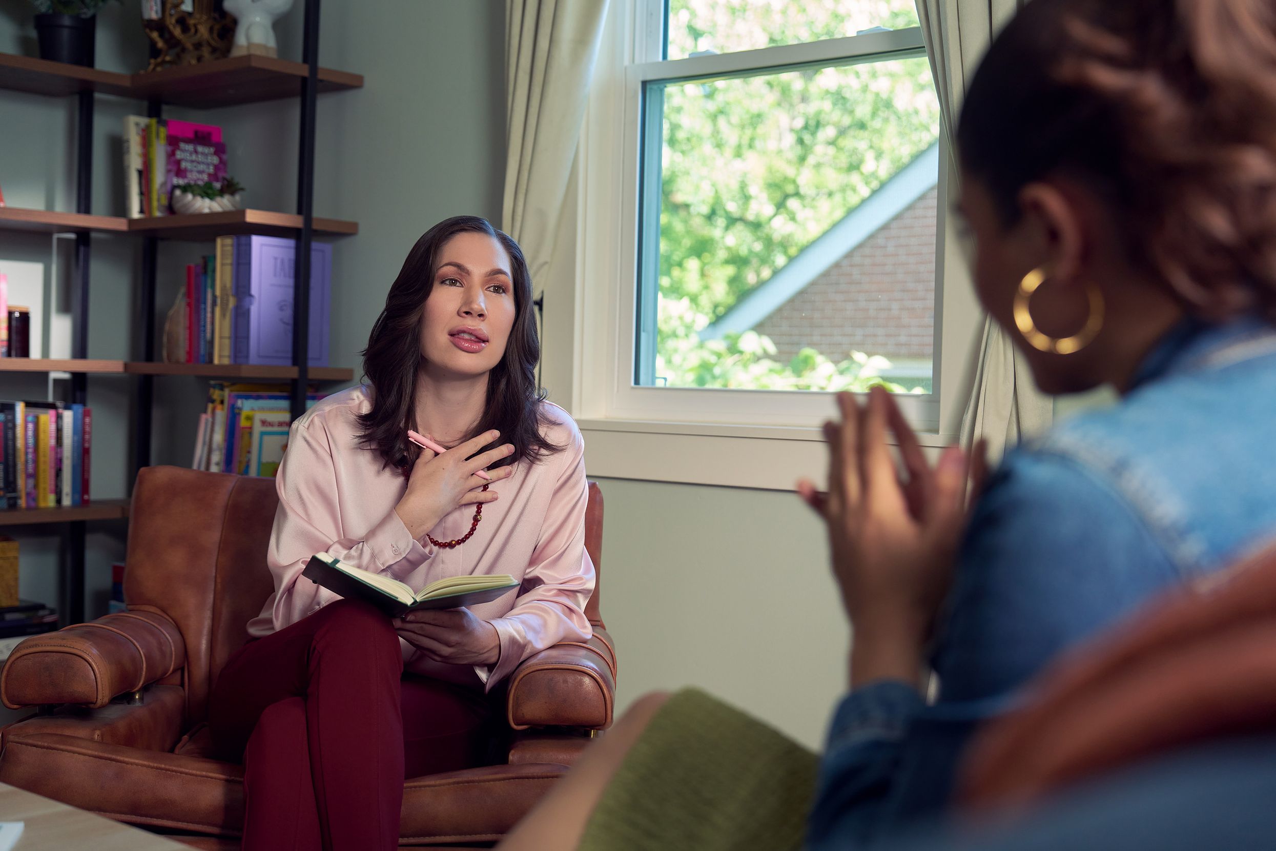 An Eastern European white woman therapist sits in a large chair and looks at a client seated on a couch in front of her. She is holding a pen and open notebook. The client is a Latina woman with dark brown wavy hair pulled into a ponytail. The therapist's office has several books on display and a window letting in natural light. The therapist is wearing a silk pink top, maroon dress pants, and a minimalist necklace. The client is wearing gold hoop earrings, a jean jacket, and green skirt.