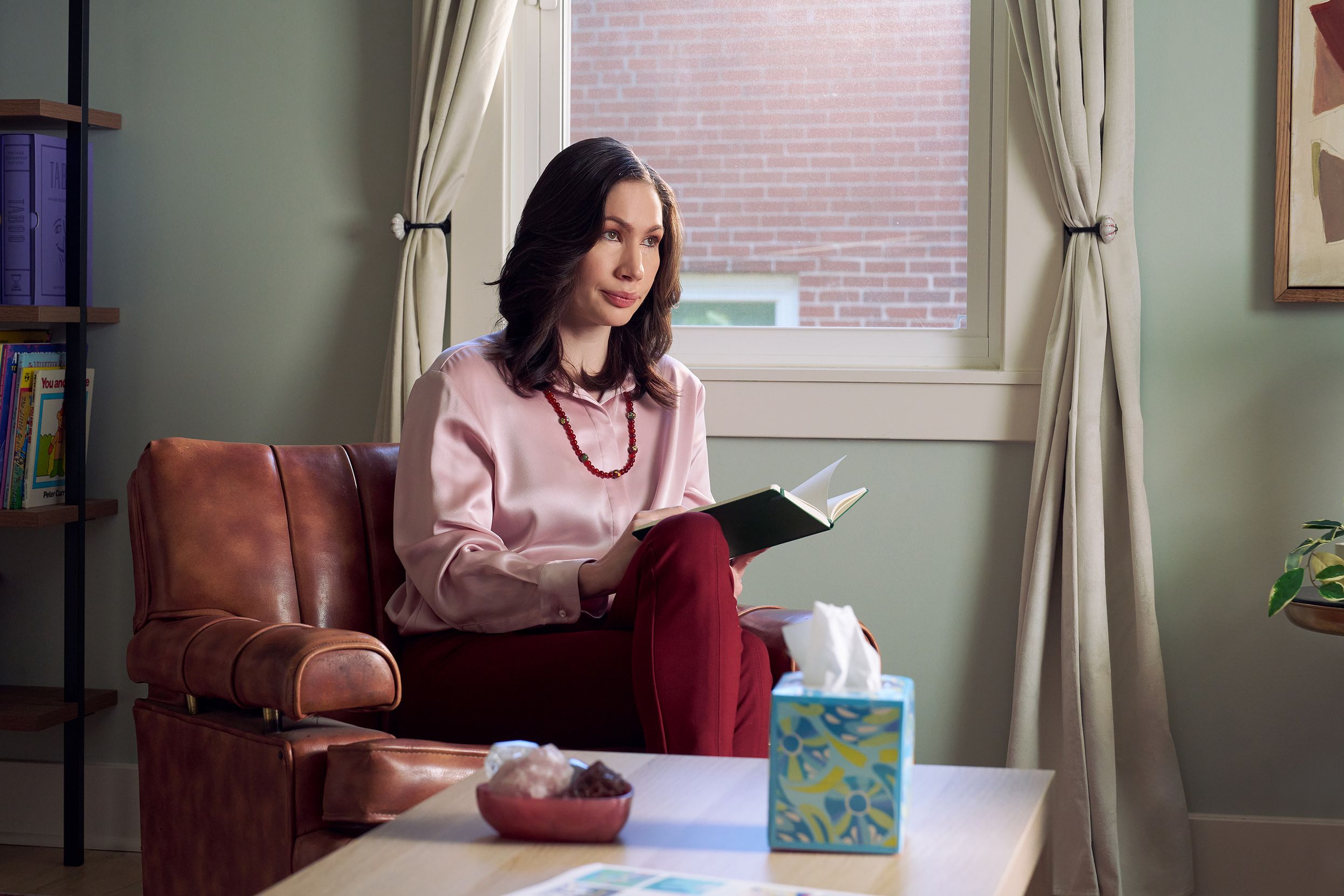 An Eastern European white woman therapist sits in a large chair looks at a client off camera. She is holding an open notebook. A tissue box and a red marble heasrt-shaped bowl of crystals are on a low coffee table in front of her. The therapist's office has several books on display and a window letting in natural light. The therapist is wearing a silk pink top, maroon dress pants, and a minimalist necklace.