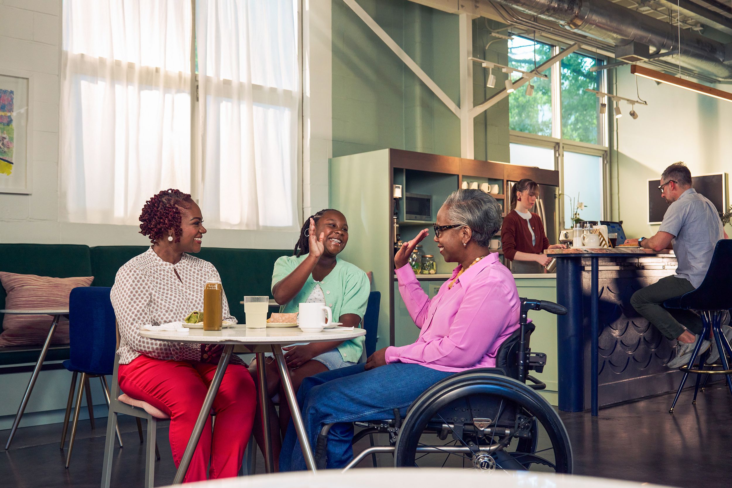 An African American daughter and auntie give each other a high five at a table in a modern cafe, while the girl's mother smiles and watches. The auntie is paraplegic and is seated in a manual wheelchair. She has short gray hair and is wearing a lavender silk blouse and dark blue jeans. The mother has short tightly curled red hair and is wearing a patterned blouse and red pants. The daughter is wearing a cochlear implant and has her braided hair pulled back into a pony tail. She is wearing a light green cardigan and jean shorts. Behind them, a barista prepares drinks and a man types on a laptop.