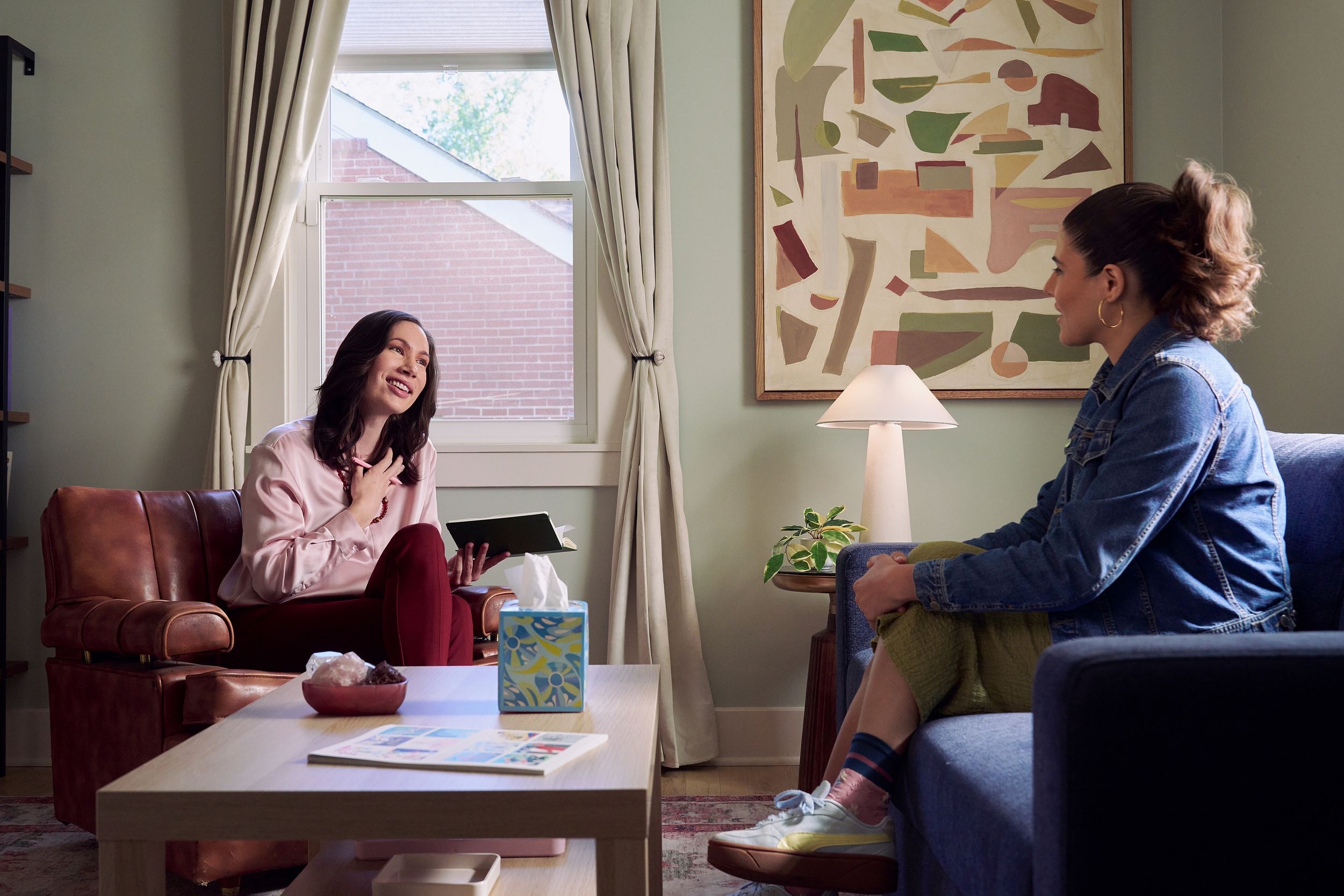 An Eastern European white woman therapist sits in a large chair and looks at a client seated on a couch in front of her. She is holding her hand to her chest and smiles at her client knowingly. The client is a Latina woman with dark brown wavy hair pulled into a ponytail. A tissue box, a red marble heasrt-shaped bowl of crystals, and a magazine are on a low coffee table in the center of the office. An abstract art piece, a lamp, and a small house plant are next and a window letting in natural light. The therapist is wearing a silk pink top, maroon dress pants, and a minimalist necklace. The client is wearing gold hoop earrings, a jean jacket, green skirt, and sneakers.
