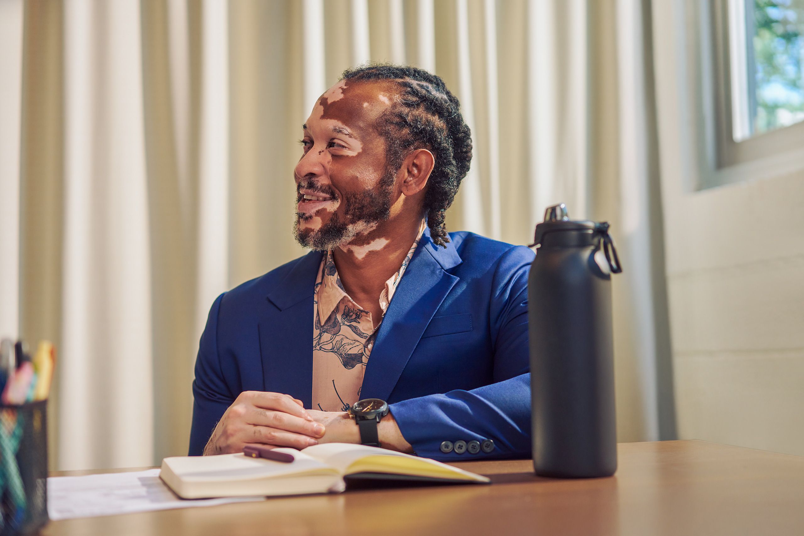 An African American man with olive and porcelain skin and Non-Segmental Vitiligo is seated at a conference room table, smiling at a colleague off-camera. A black water bottle is in front of him. He has brown barrel-twist dreadlocks, wears a navy blue suit with light pink and floral blue-patterned dress shirt. His brown eyes and black watch offset his black goatee.