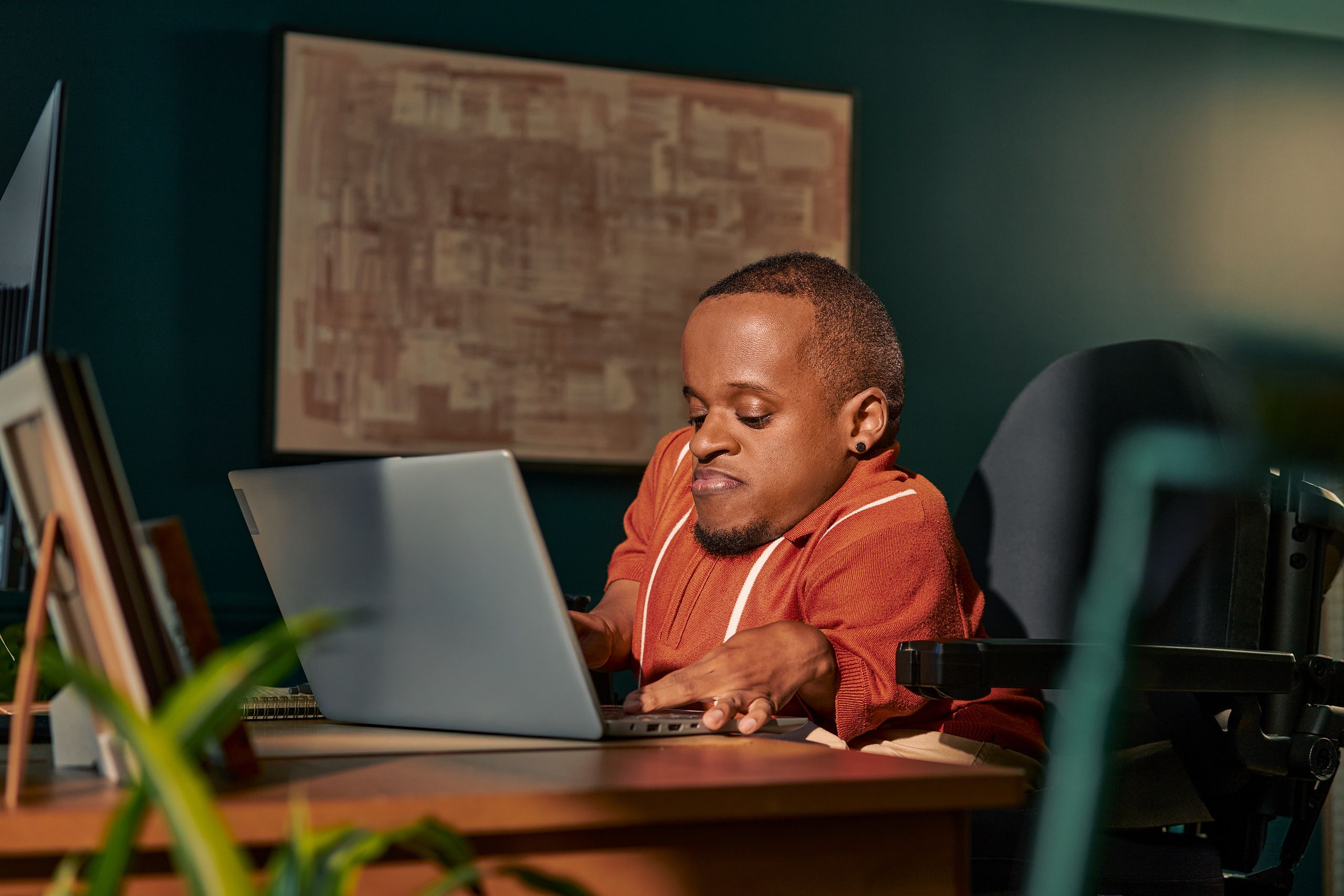 A black man who is a little person is seated in his power wheelchair in a dark teal home office. He types on a laptop on his desk, with a monitor and framed photos nearby. An abstract art piece is on the wall behind him. He has closely cropped black hair, brown eyes, and is wearing a rust-orange knit quarter-zip sweater with white vertical stripes, light khaki trousers, and a small stud earring.