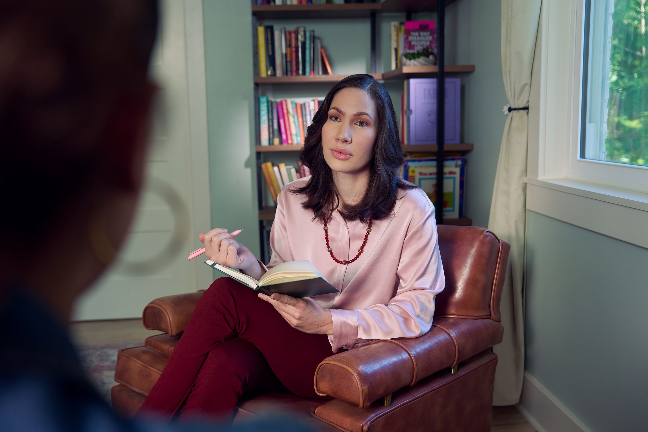 An Eastern European white woman therapist sits in a large chair with an understanding look on her face. She is holding a pen and open notebook, and is looking at a client off camera. The therapist's office has several books on display and a window letting in natural light. The therapist is wearing a silk pink top, maroon dress pants, and a minimalist necklace.