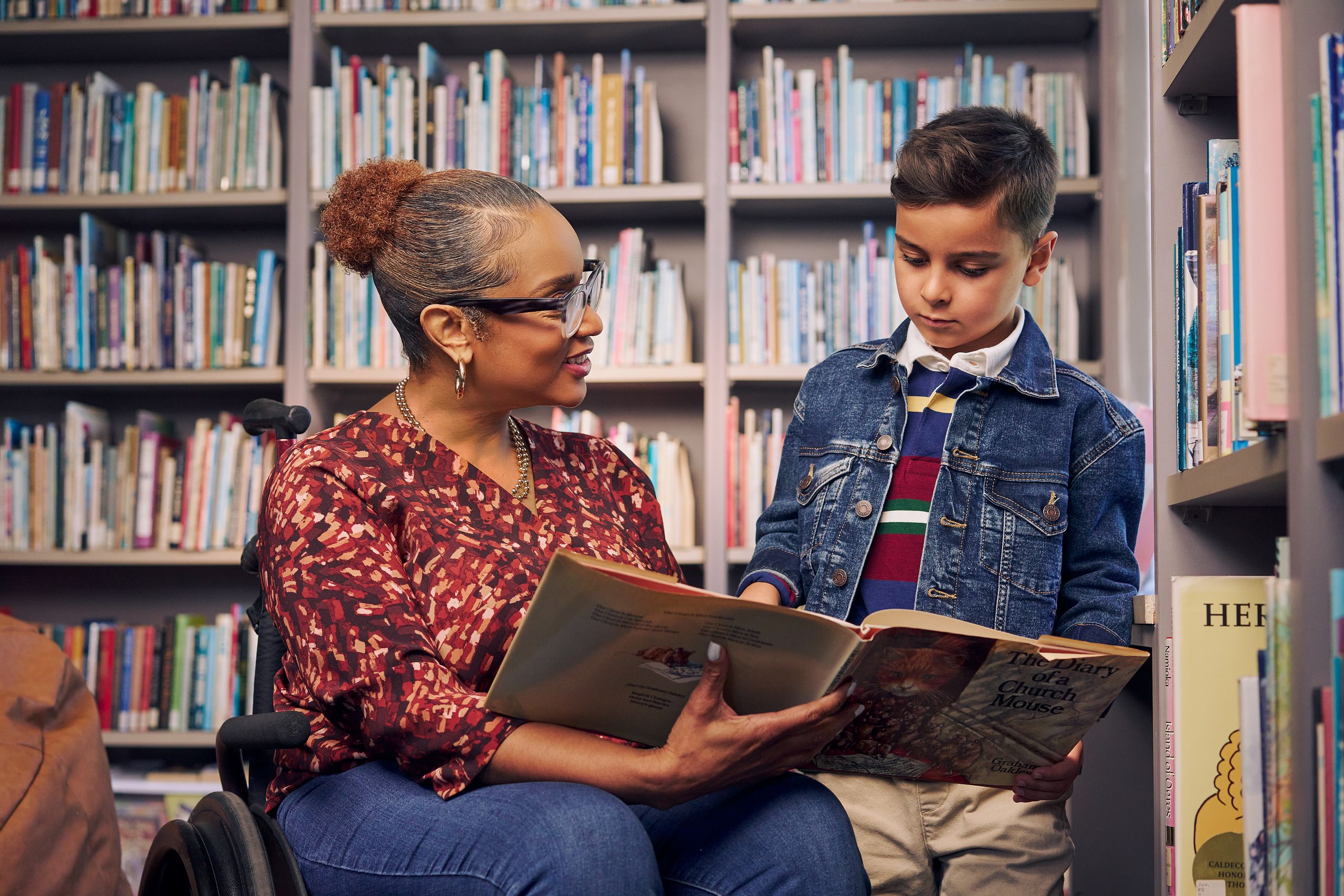 An African American teacher seated in a manual wheelchair holds a picture book open while her young student reads out loud. The teacher has medium brown skin, reddish brown hair pulled into a short natural ponytail, and is wearing reading glasses, a red patterned blouse, and jeans. The young student is a Hispanic and Mediterranean-American boy with short brown hair. He has olive skin and is wearing a blue striped shirt, a jean jacket, and khaki pants. A wall of children's books are behind them.