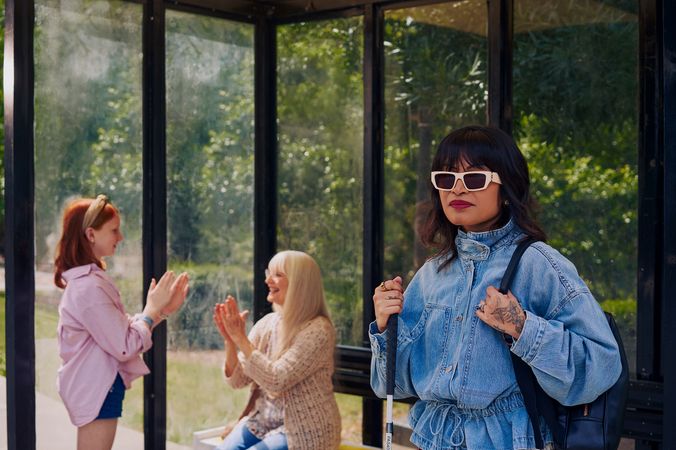 An Asian Filipino woman with Retinitis Pigmentosa and Usher Syndrome stands at a covered bus stop. She is wearing white sunglasses and is holding her white cane with her tattooed hands. She has on a jean jacket and a black backpack. Behind her, a white European girl with red hair plays pattycake hand game with her grandmother, who is sitting on the bus stop bench. Behind the bus stop are several large trees and walking paths.