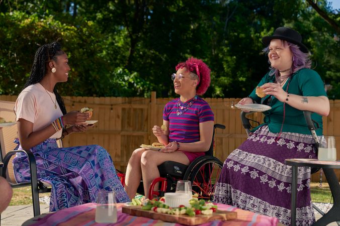 At a backyard barbeque, two nonbinary people sit in their mobility aids, chat with a friend while holding plates of food.  One nonbinary person, a Black person with a curly pink fro with loc bangs wearing glasses is seated in a manual wheelchair. They are wearing bright pink sunglassese on their head, a purple and pink striped crop top, pink shorts, and purple sandals. They have several tattoos on both legs. The second nonbinary person, a white person with layered purple hair with two long thin red braids on each side, is seated on a dark gray rollator. They are wearing a black hat, a green shirt, and a purple patterned skirt. Their friend, an African American woman with long braided hair, is wearing a pink shirt and patterned blue and purple skirt.