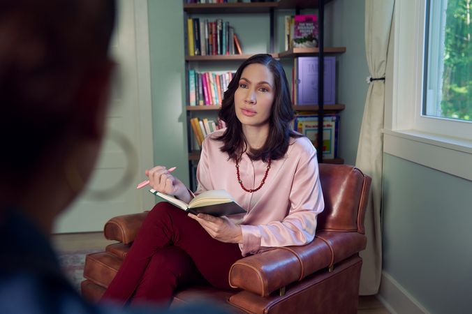 An Eastern European white woman therapist sits in a large chair with an understanding look on her face. She is holding a pen and open notebook, and is looking at a client off camera. The therapist's office has several books on display and a window letting in natural light. The therapist is wearing a silk pink top, maroon dress pants, and a minimalist necklace.